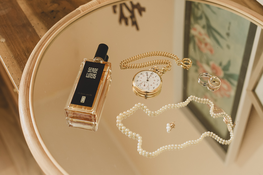 parfum, bijoux, montre à gousset et alliances, photo des détails mariage posés sur un miroir