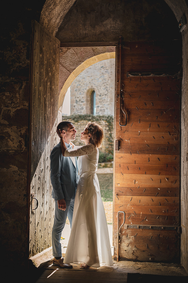 Couple face à face dans l’entrebâillement d'une grande porte en bois de l'abbaye de Beauport et la lumière rend la robe de mariée légèrement transparente