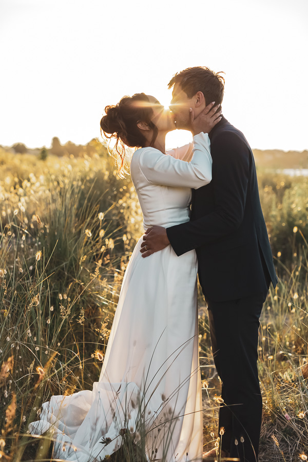 Les mariés s'embrassent au coucher du soleil dans une prairie avec des queues de lièvres devant la mer