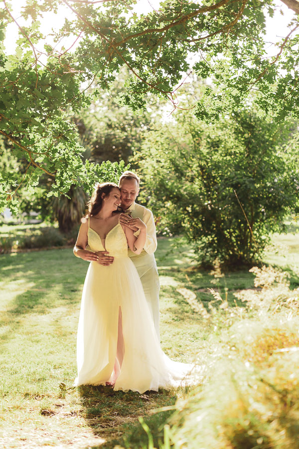 Couple de mariés dans les jardins du lieu de réception "L'eau des collines" sous les arbres avec la lumières en contrejour
