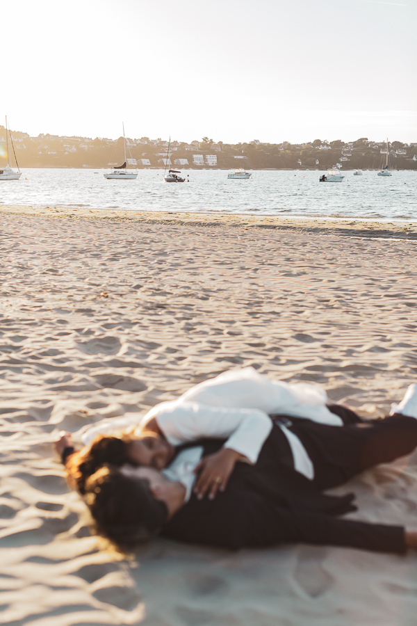les mariés alongés et enlacés sur la plage avec les bateaux en arrière plan au coucher de soleil