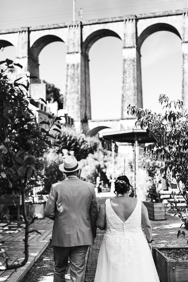 Les mariés vue de dos marchent dans les jardins devant la mairie. Le viaduc en arrière plan