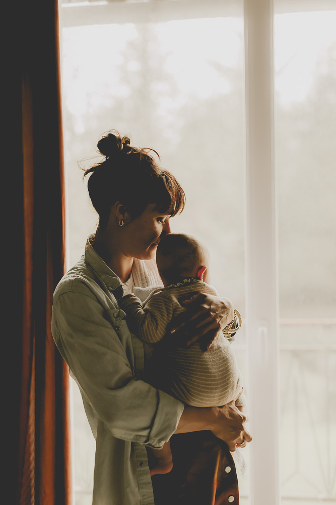 maman porte bébé dans ses bras devant la fenetre contre jour